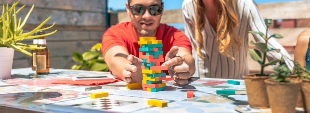 Concentrated young man with sunglasses pushing jenga game piece next to his female friend in rooftop on a summer party.