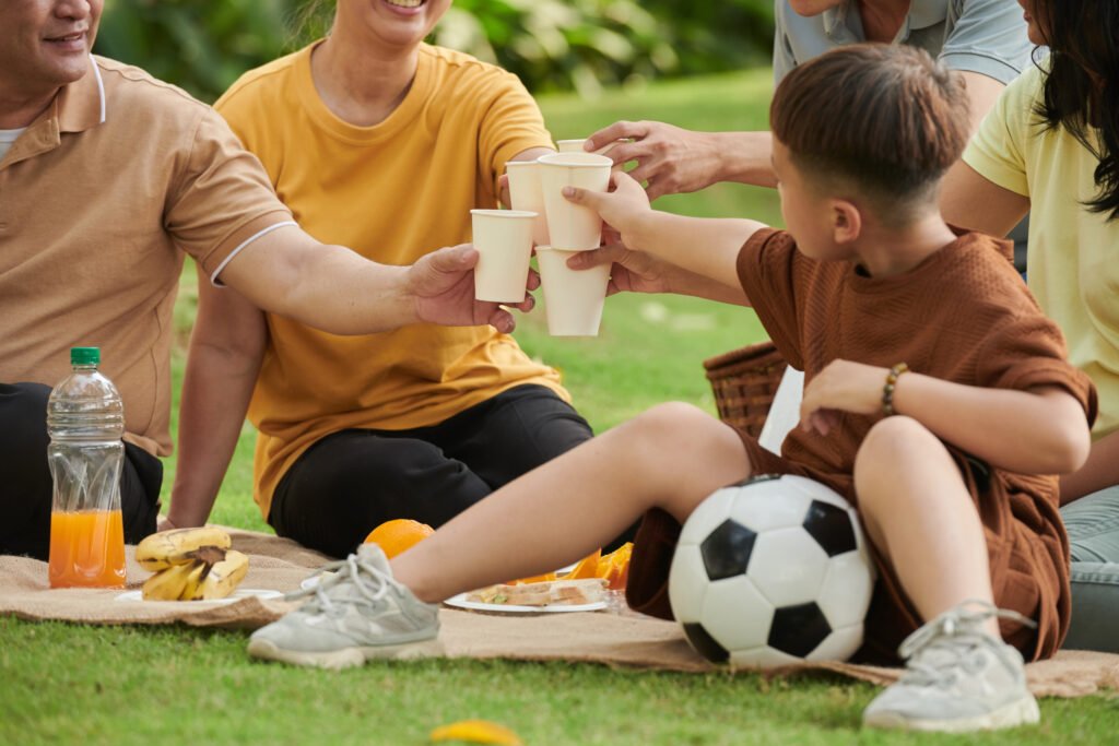 Family toasting with paper cups with refreshng drinks when enjoying picnic in green environmental park.