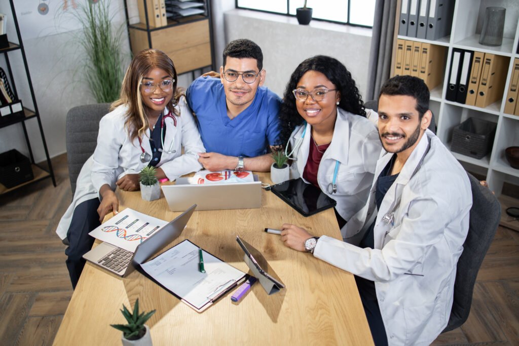 Group of doctors wearing coats and scrubs sitting together at table and smiling on camera. Medical specialists during session.