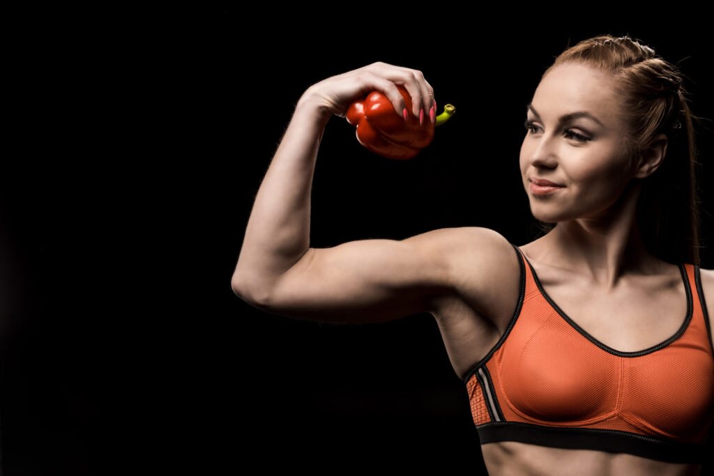 smiling sporty girl holding bell pepper solated on black.