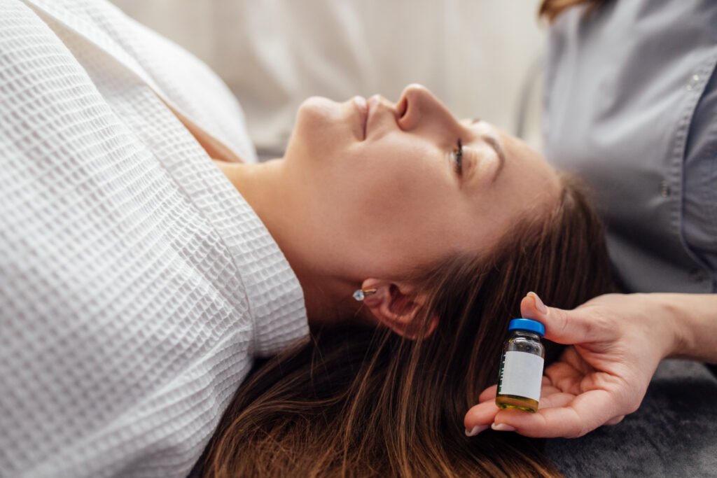 A close-up of a cosmetologist hand with a mock-up of a glass bottle with peeling liquid. A young female client in a bathrobe is lying 