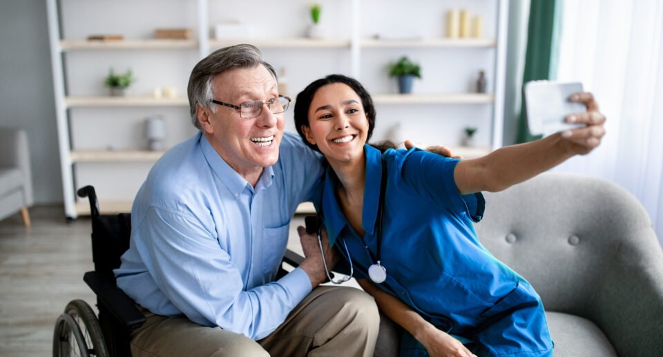 Cheerful impaired senior man in wheelchair taking selfie with young female doctor at retirement home. Disabled older patient making photo with nurse, having fun together indoors. concept of home health care.