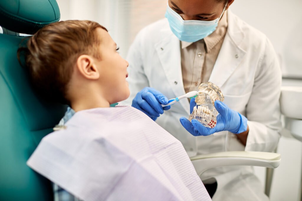 African American female dentist using dentures while teaching kid how to brush his teeth properly at dental clinic.