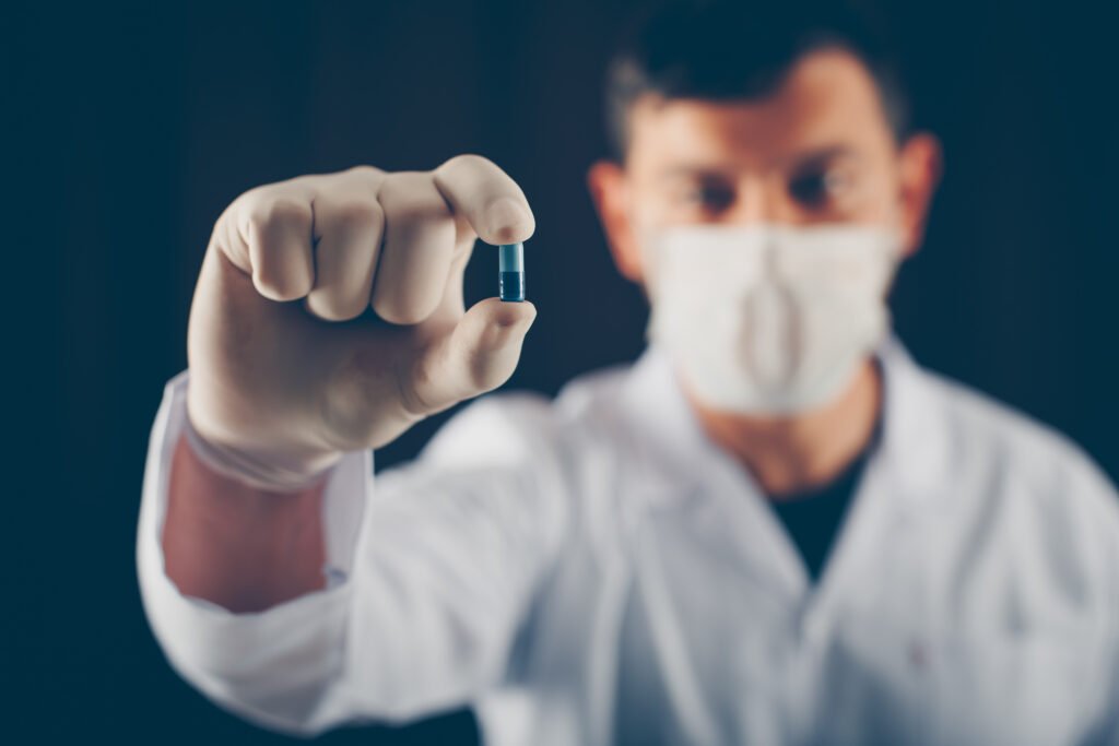 Doctor with mask and gloves holding blue pill with his fingers on a dark background. side view.