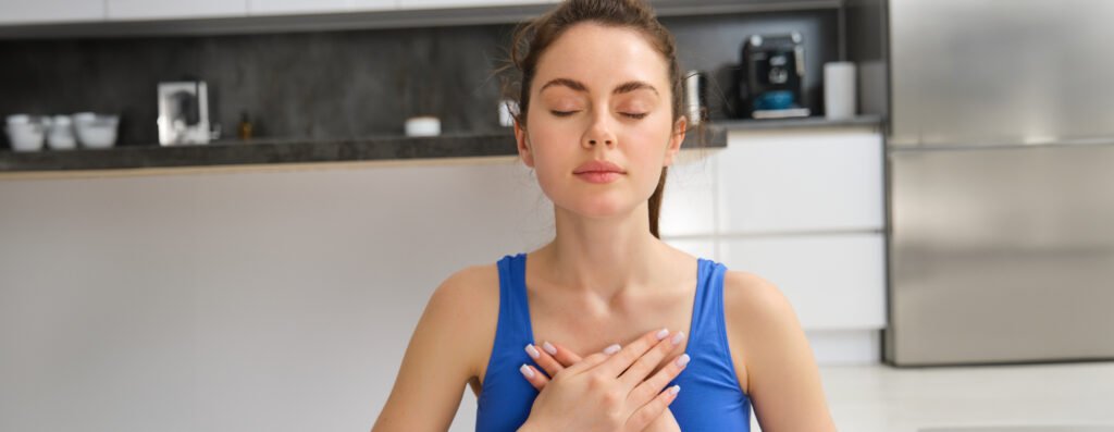 Image of calm and relaxed woman meditating, doing breathing practices, holding hands on chest during yoga session at home. Sport and mindfulness concept.