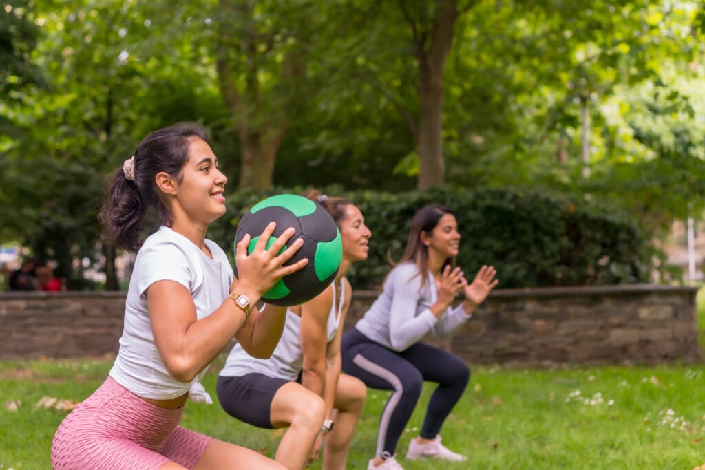 Latin girl doing sports in a green park, lifestyle a healthy life, teacher with the students doing squats with the weight ball