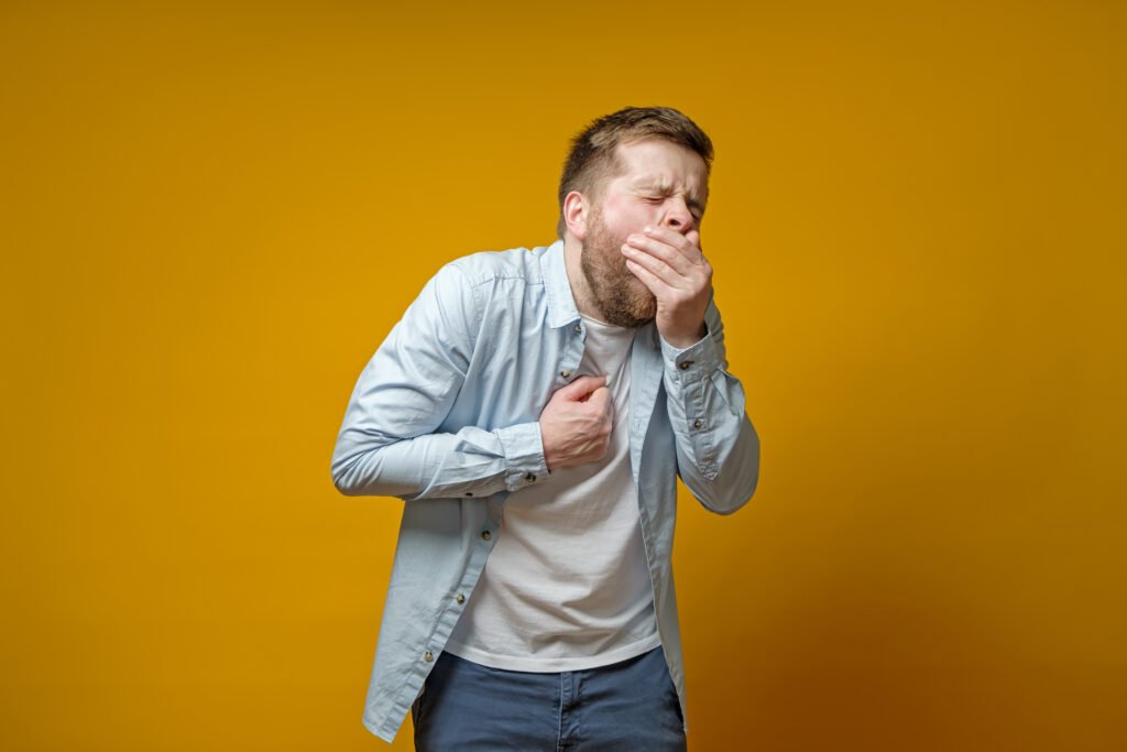 Bearded man coughs, covering mouth with palm and holds hand on chest, experiencing pain. Concept of the shortness of breath.