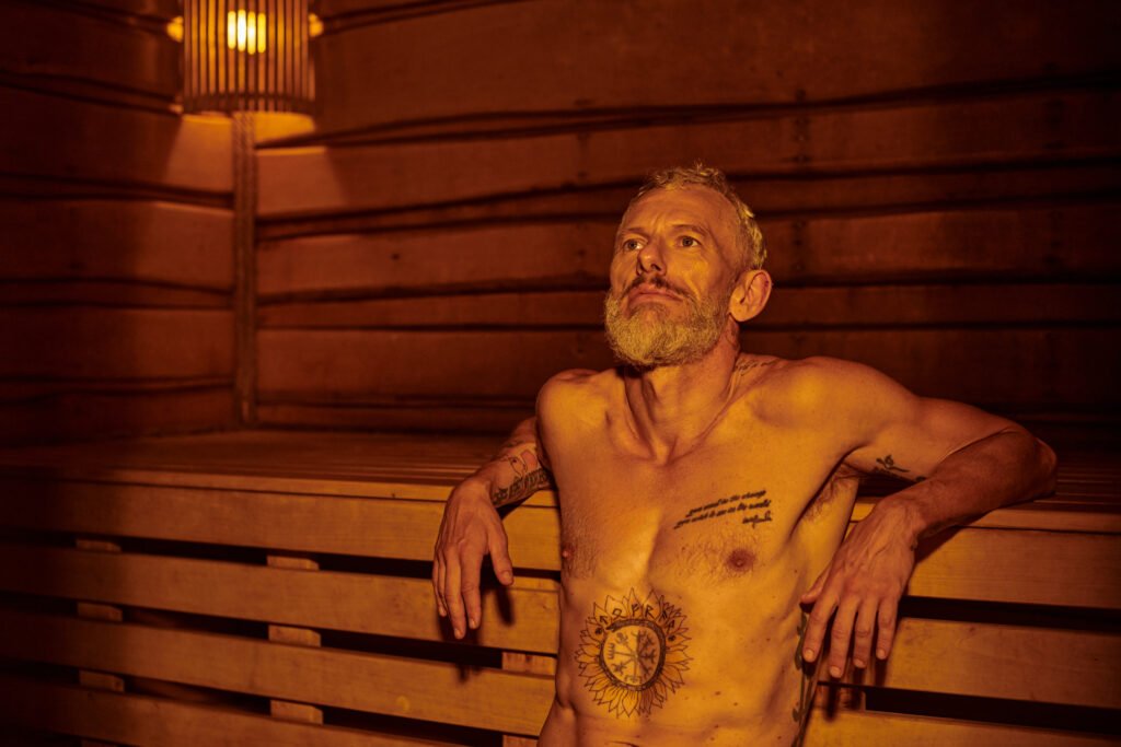 pensive and shirtless middle aged man with tattoos sitting in sauna, wellness retreat concept