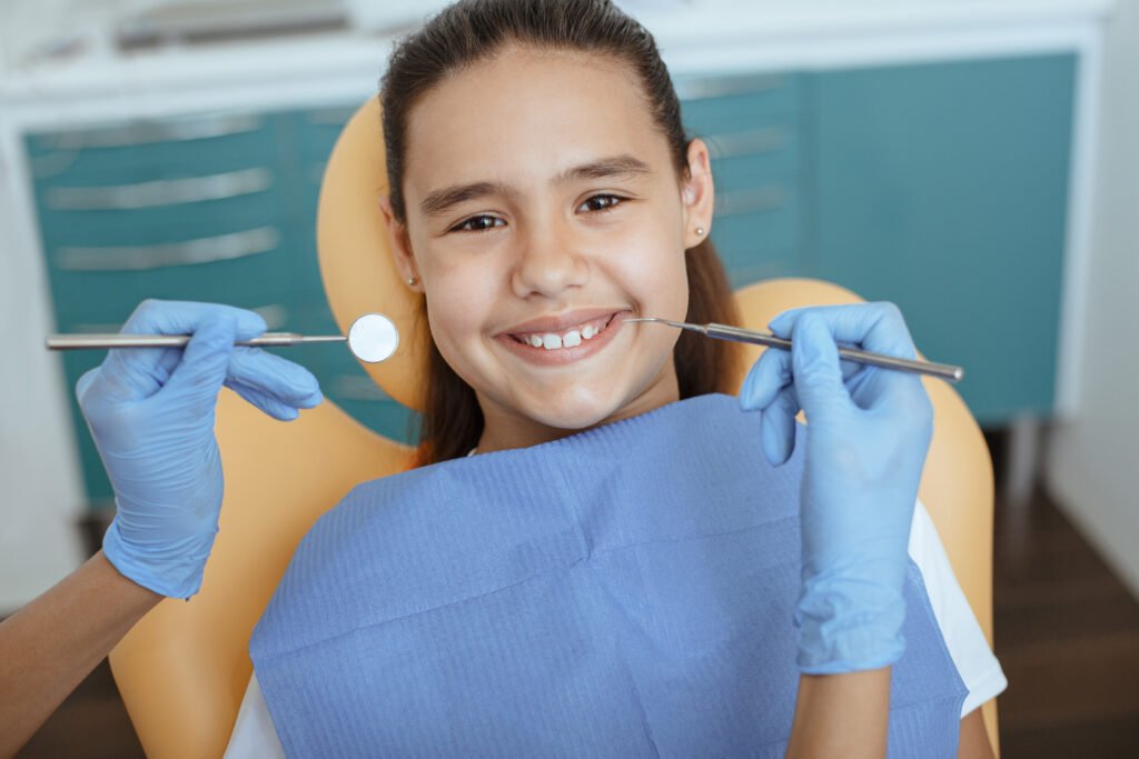 Prevention and treatment of teeth in children. Cheerful cute little girl sit in medical chair and look at camera, woman doctor in rubber gloves with tool begins procedure in interior of modern clinic