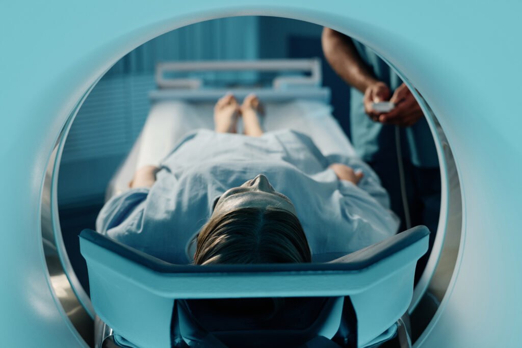 Mature Caucasian woman in blue gown lying on CT scanner bed in modern clinic, unrecognizable doctor adjusting settings