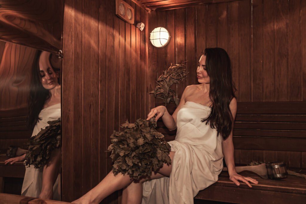 Russian bathhouse. Adult woman relaxing and sweating in wooden sauna with hot steam. Female with bath besom resting on bench in spa complex. Wellness, self care, healthy concept.