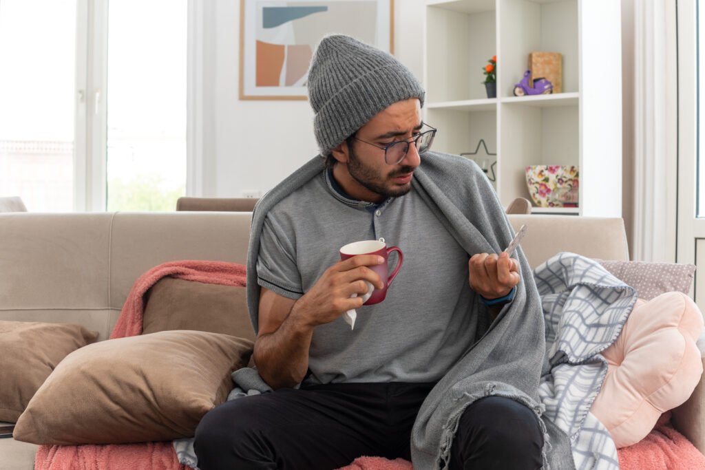 sick young caucasian man in optical glasses wrapped in plaid wearing winter hat holding cup and looking at medicine blister pack sitting on couch at living room