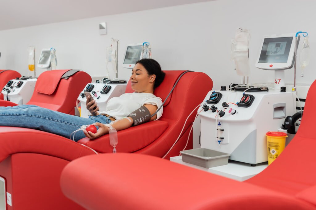 smiling multiracial woman with rubber ball and transfusion set browsing internet on smartphone while sitting on medical chair near automated equipment during blood donation in clinic