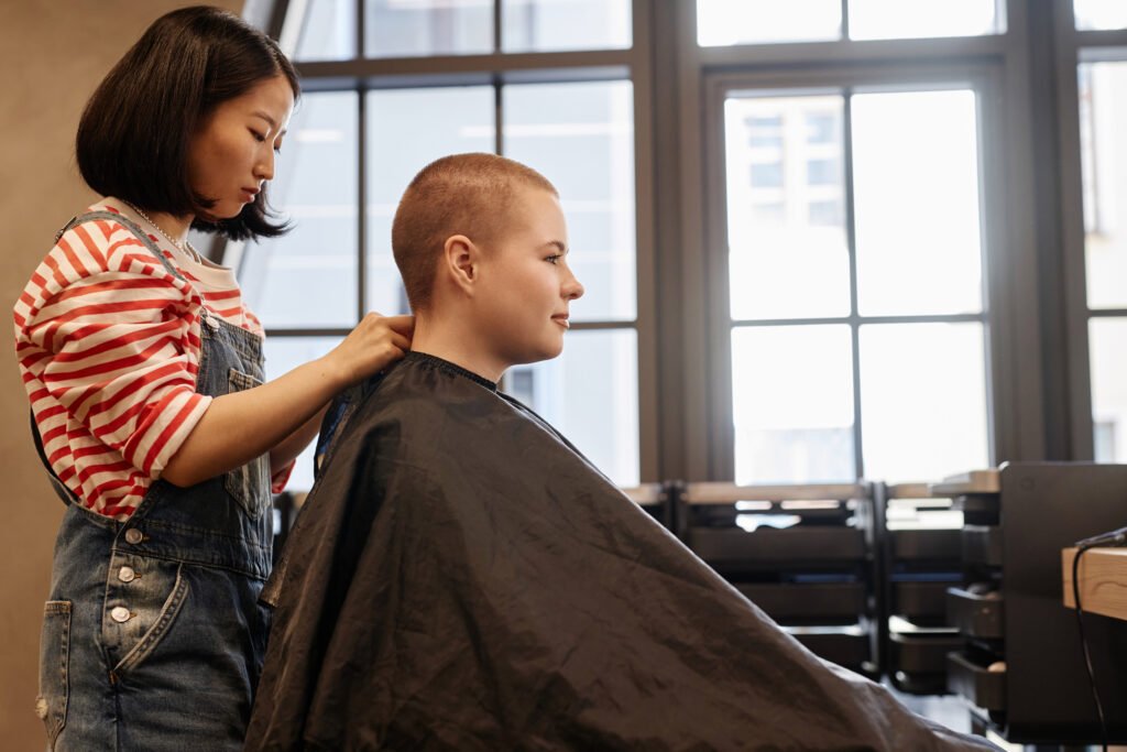 Side view portrait of smiling young woman with buzzcut sitting in salon chair with hairstylist working