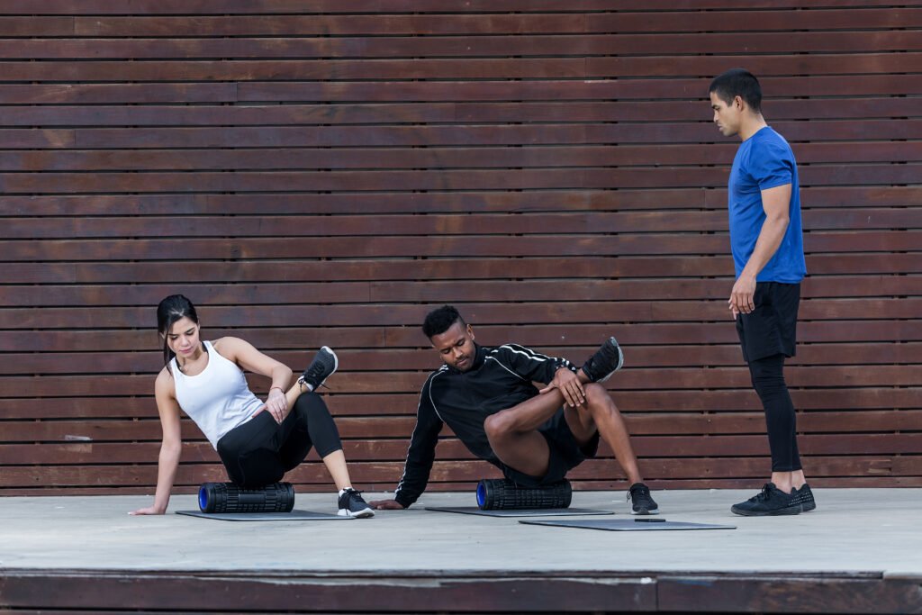 Adult personal instructor standing near wooden wall and looking at multiethnic man and woman performing exercise on rubber roller on city street.