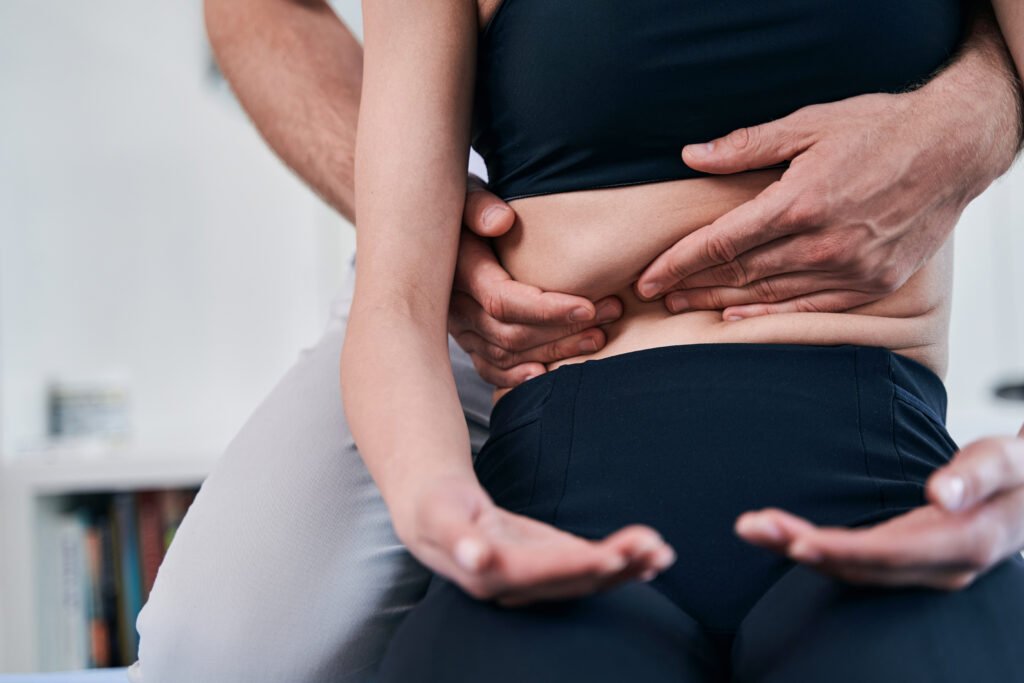 Young woman in black sportswear sitting on medical couch while getting professional care on her stomach in wellness center to improve shortness of breath.