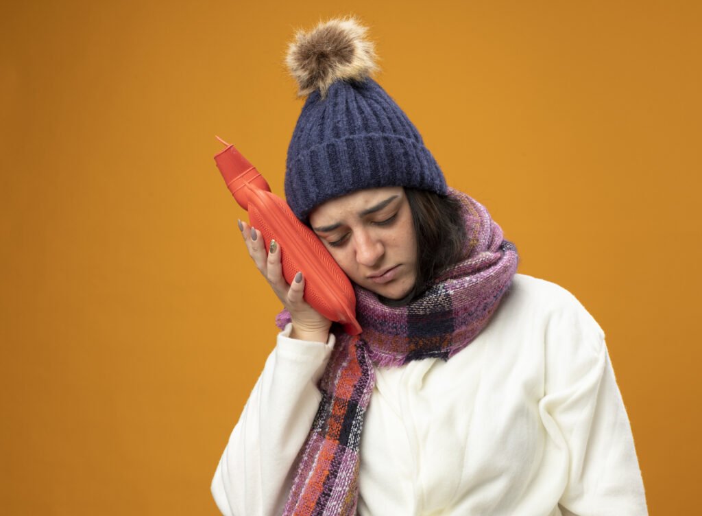 weak young caucasian ill girl wearing robe winter hat and scarf touching head with hot water bag with closed eyes isolated on orange background with copy space.