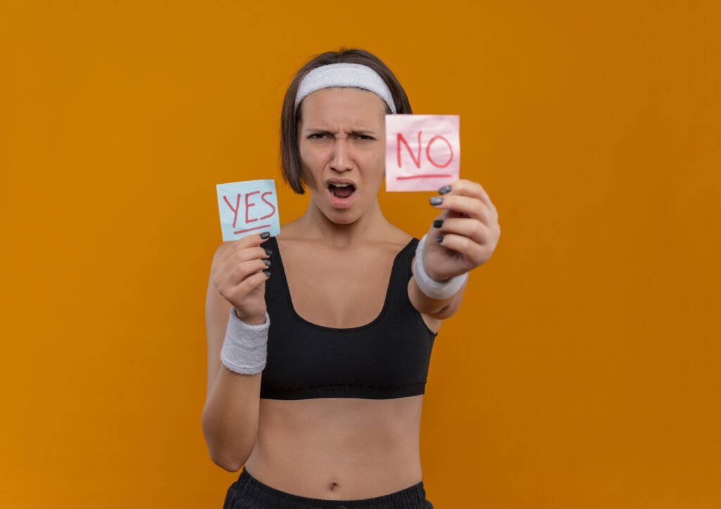 young fitness girl in sportswear with headband showing two reminder papers with word yes and no smiling cheerfully standing over orange background