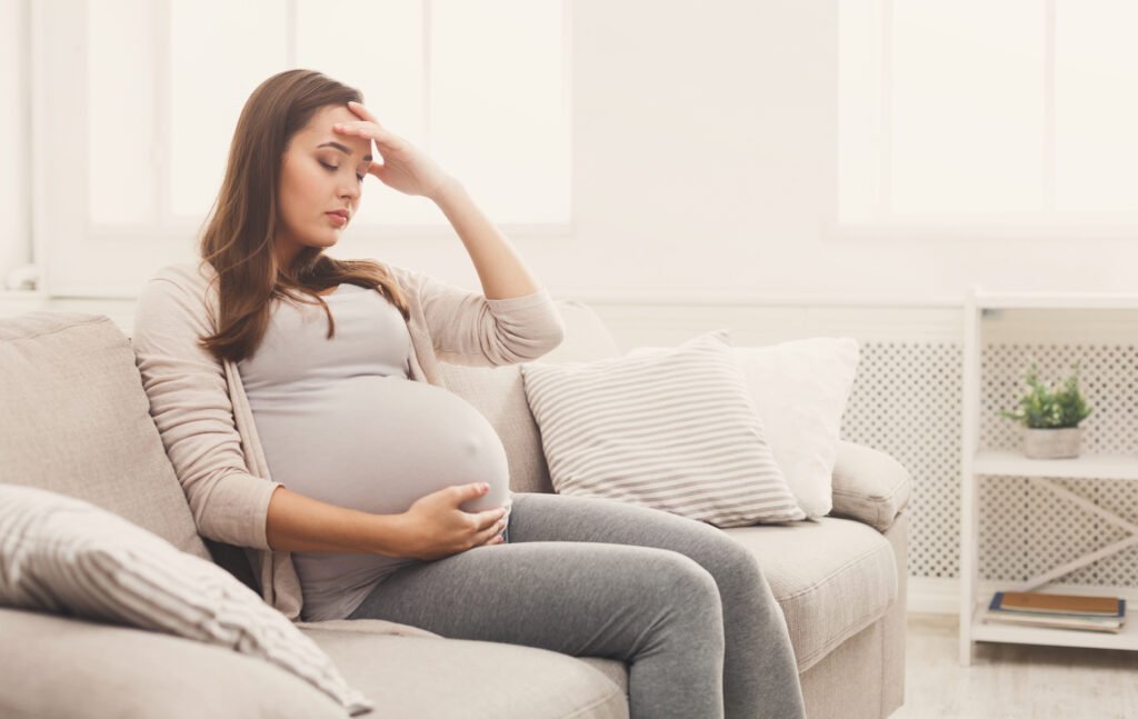 Young pregnant woman with headache sitting on sofa. Pregnancy symptoms,suffering from headich due to hormonal imbalance.