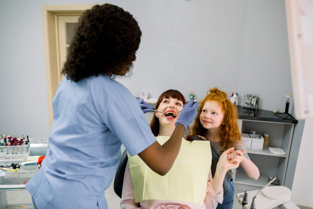 Young woman, sitting in dental chair, supporting by her little daughter, while black female dentist cures her teeth. Back view of dentist wearing latex gloves and holding tools in hands.