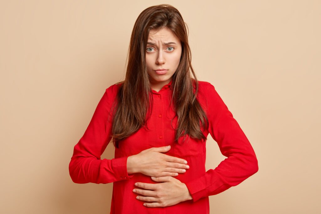 Photo of attractive dark haired woman keeps hands on stomach, suffers from hormonal imlabance, has spasm, dressed in red shirt, poses over beige background. Bad feelings concept. Female has menstruation